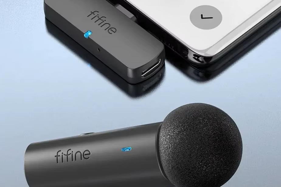 Microfone de lapela sem fio Fifine em primeiro plano sobre uma superfície refletora, com o recetor USB-C ligado a um telemóvel.
