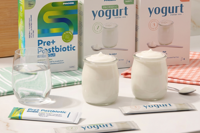 Imagem de três caixas de produtos Prozis (Pre+ Postbiotic e misturas para iogurte) numa bancada branca, acompanhadas por um copo de água, dois frascos de vidro com iogurte cremoso e três saquetas individuais.