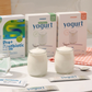 Imagem de três caixas de produtos Prozis (Pre+ Postbiotic e misturas para iogurte) numa bancada branca, acompanhadas por um copo de água, dois frascos de vidro com iogurte cremoso e três saquetas individuais.