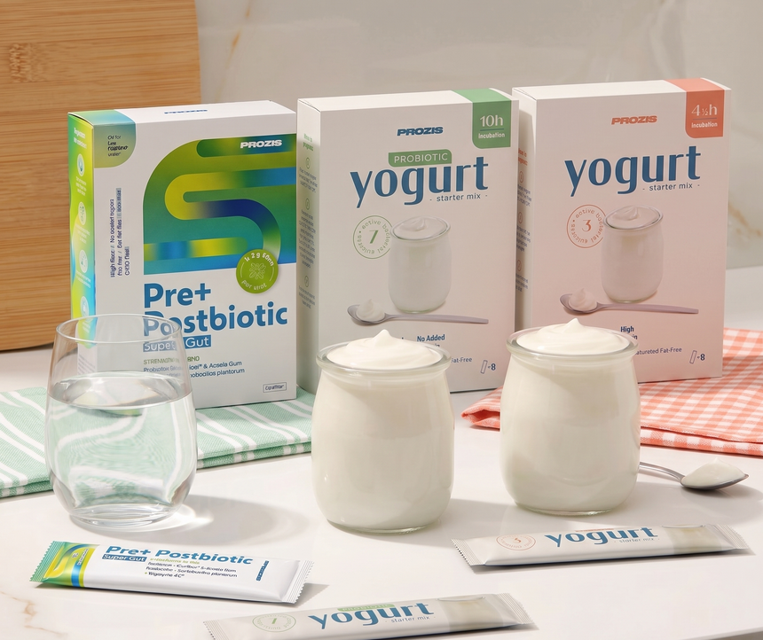 Imagem de três caixas de produtos Prozis (Pre+ Postbiotic e misturas para iogurte) numa bancada branca, acompanhadas por um copo de água, dois frascos de vidro com iogurte cremoso e três saquetas individuais.