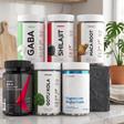 Seis produtos PROZIS organizados em duas filas sobre uma bancada de mármore. A fila de trás mostra os suplementos GABA, Shilajit e Maca Root. A fila da frente mostra EAA, Gotu Kola e Magnesium Bisglycinate. O fundo é um ambiente de cozinha.