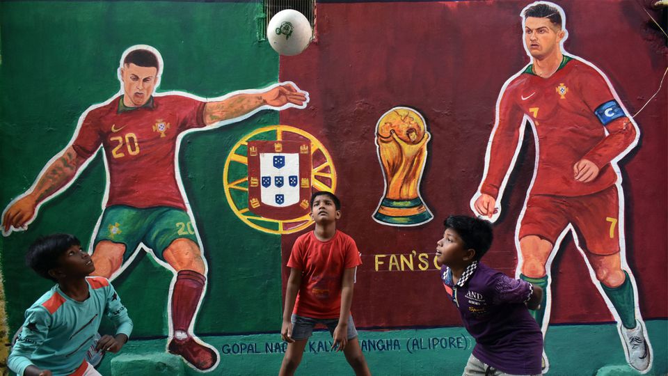 Crianças brincam com uma bola em Calcutá, na Índia, em frente a mural com as figuras de Cristiano Ronaldo e João Cancelo