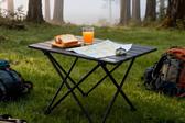 Mesa de campismo portátil em alumínio com lanche, mapa e bússola, montada num cenário de natureza com árvores e neblina.