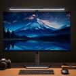 Luz magnética de ecrã Baseus i-wok3 montada num monitor. A luz ilumina uma mesa de trabalho em madeira com teclado, rato, caderno e câmara.