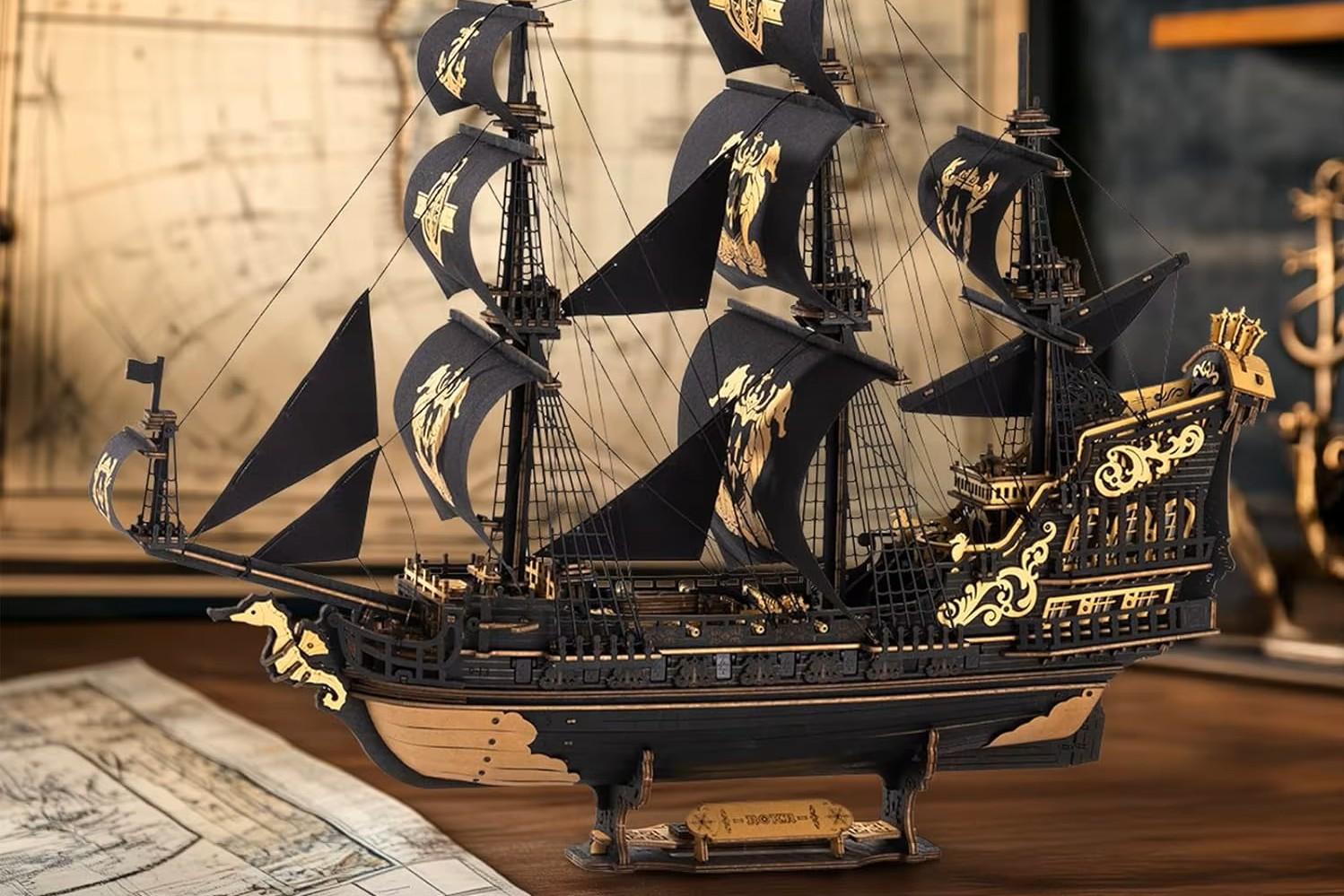 Modelo 3D de um barco. A maquete está apoiada sobre uma mesa de madeira, com mapas antigos compondo o cenário ao fundo.
