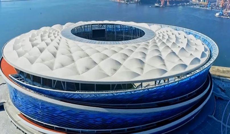 Estádio Dalian Suoyuwan – Dalian, China. Construído à beira-mar, inspira-se em temas marítimos — ondas, conchas e tons de azul oceânico. Uma rampa em dupla hélice envolve a fachada em espiral Foto: Youtube