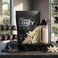Embalagem de Prozis Supreme Whey Protein num ginásio, acompanhada por um shaker cheio, uma toalha de treino e flores de baunilha sobre uma base de pedra.