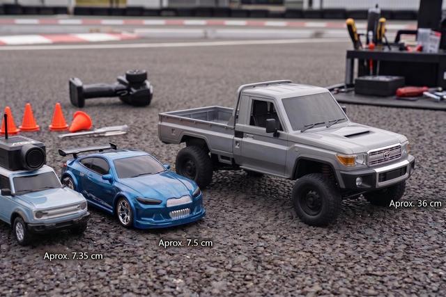 Três carros de controlo remoto de diferentes modelos e tamanhos, alinhados lado a lado numa superfície de asfalto. No fundo, desfocados, estão pequenos cones de trânsito, um comando à distância e algumas ferramentas.