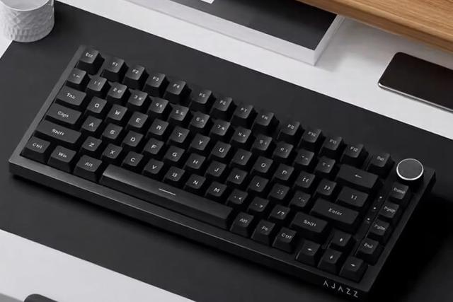 Teclado mecânico da marca AJAZZ, com um botão giratório no canto superior direito, posicionado sobre um tapete numa secretária.