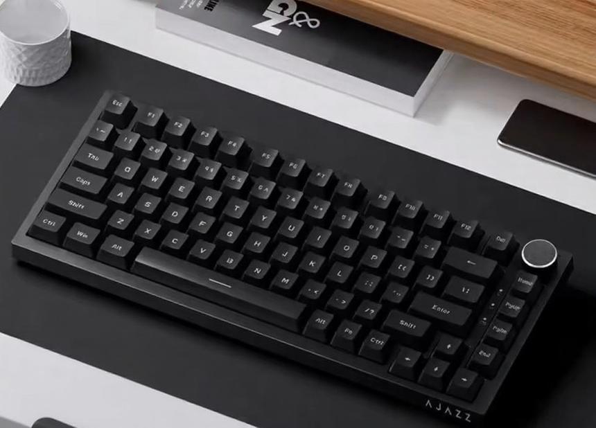 Teclado mecânico da marca AJAZZ, com um botão giratório no canto superior direito, posicionado sobre um tapete numa secretária.