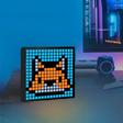 Um ecrã digital sobre uma secretária exibe uma raposa em pixel art. Ao fundo, num ambiente iluminado, um monitor, um pequeno dispositivo com luz azul, um tablet e um teclado.