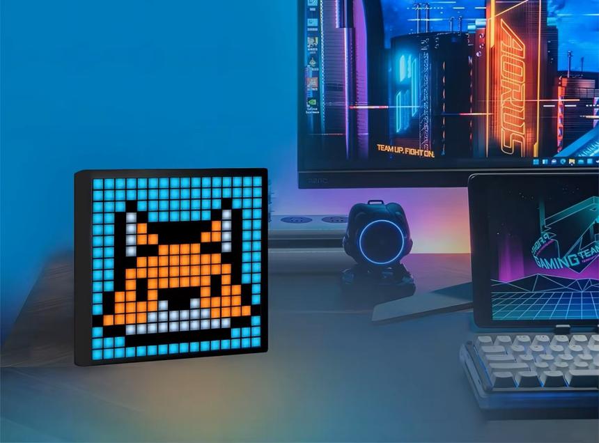 Um ecrã digital sobre uma secretária exibe uma raposa em pixel art. Ao fundo, num ambiente iluminado, um monitor, um pequeno dispositivo com luz azul, um tablet e um teclado.