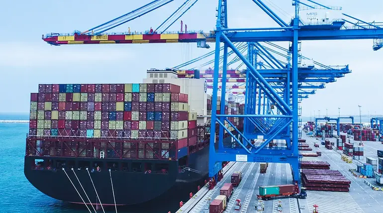 Large container ship docked under blue cranes at a modern West African port terminal.