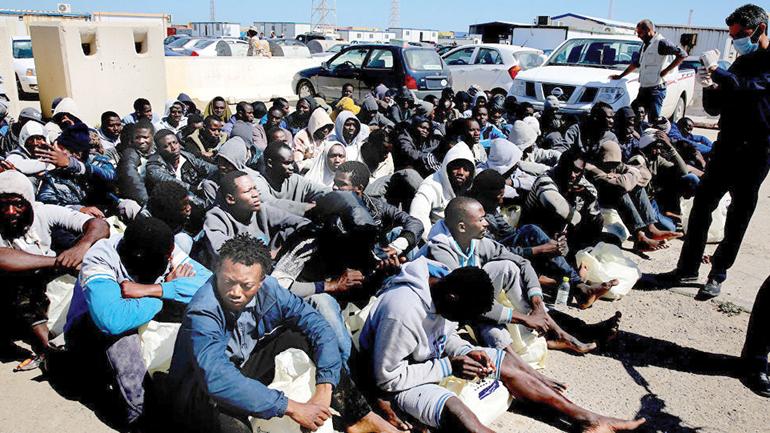 A large group of African migrants sitting on the ground outdoors at a transit or detention camp in Libya.