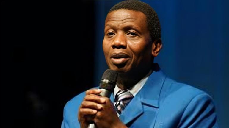 A high-resolution portrait of RCCG General Overseer Pastor Enoch Adeboye in a blue blazer and striped tie, holding a microphone with clasped hands, speaking on a stage with blurred audience lighting.