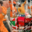 Nigerian ship chandlers in orange overalls loading fresh food crates and supplies onto a vessel.