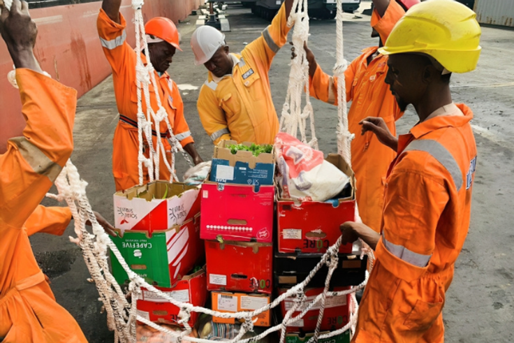 Nigerian ship chandlers in orange overalls loading fresh food crates and supplies onto a vessel.