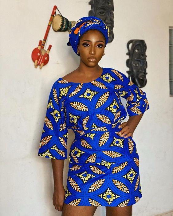 A woman posing in a short, vibrant blue and yellow pattern Ankara Oleku outfit with a matching headwrap, standing in front of a white wall adorned with traditional African masks and decorative instruments.