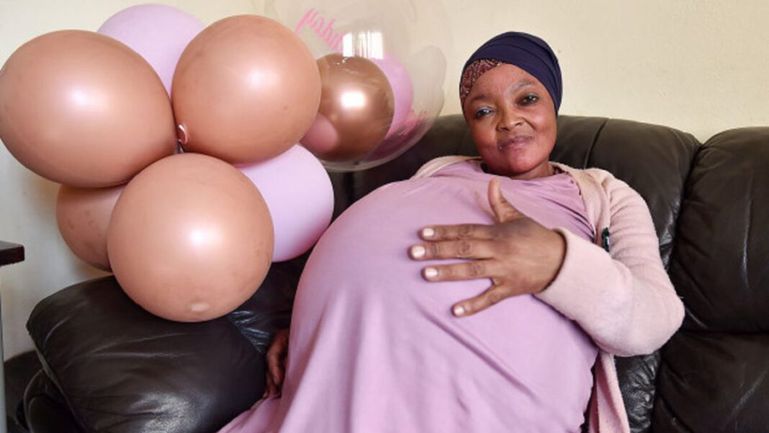 alima Cisse smiling while sitting on a sofa, heavily pregnant with record-breaking nonuplets and holding her belly next to pink balloons.