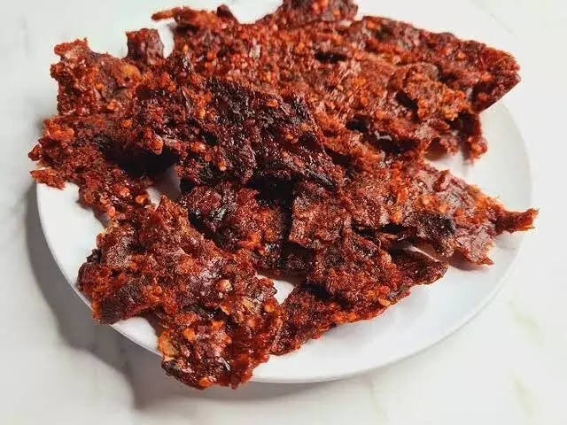 A close-up shot of a white ceramic plate piled high with pieces of dark red, spicy Nigerian kilishi (dried beef jerky) coated in pepper flakes.