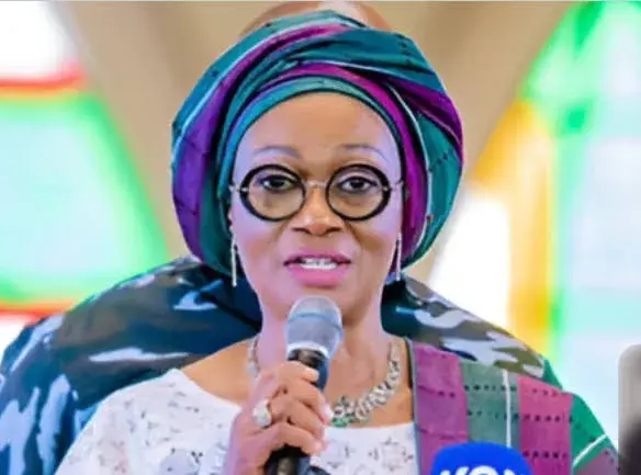 Senator Oluremi Tinubu, the First Lady of Nigeria, wearing a traditional green headtie and glasses while speaking into a microphone.