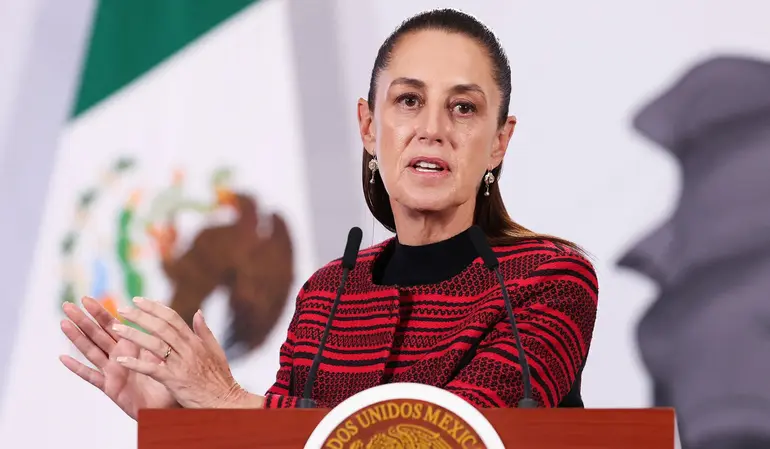 Mexican President Claudia Sheinbaum speaking at a podium with microphones, gesturing with her hands in front of the Mexican flag.