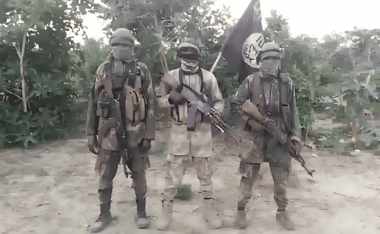 Three armed insurgents standing side-by-side in a wooded area. They are wearing full military camouflage and face masks covering their identity, holding assault rifles, with a black jihadist flag visible in the background.