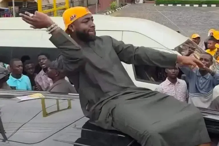 Nigerian singer Davido sitting on top of a black vehicle in a traditional green kaftan and yellow cap, gesturing to a crowd of cheering supporters.