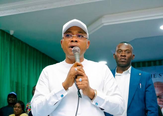 Edo State Governor Monday Okpebholo wearing a white cap and native outfit, holding a microphone and addressing a crowd at an APC rally.