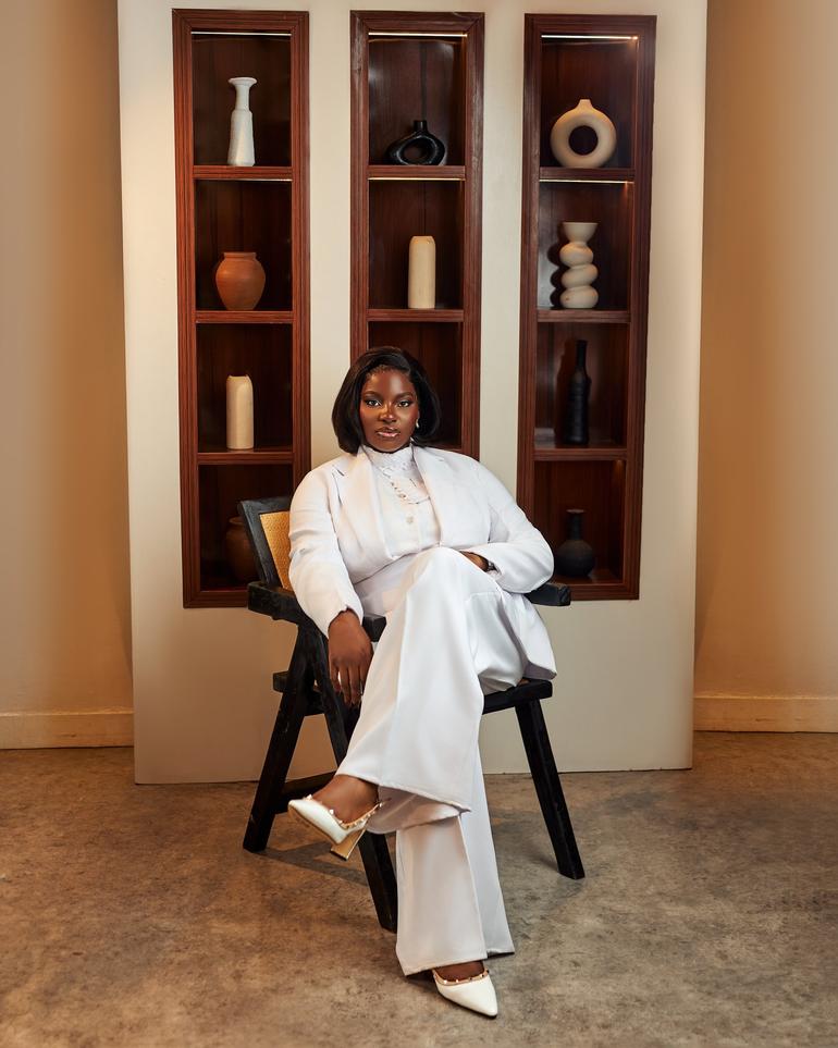 Nigerian law graduate Esther Agboola seated in a white power suit against a decorative wooden shelves background.