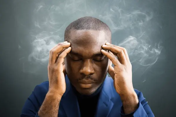 A stressed Black businessman holding his hands to his temples with his eyes closed as smoke dramatically rises around his head.
