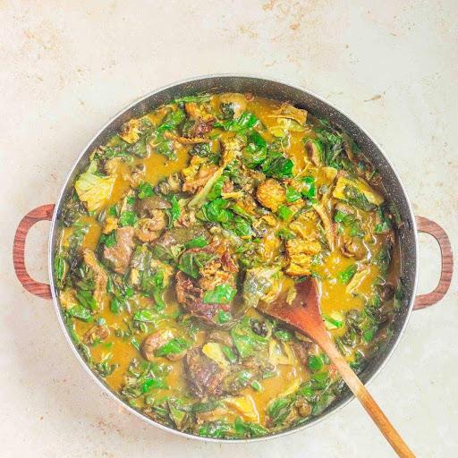 A large pot of Ofe Oha soup with green leaves and meat, with a wooden spoon inside.