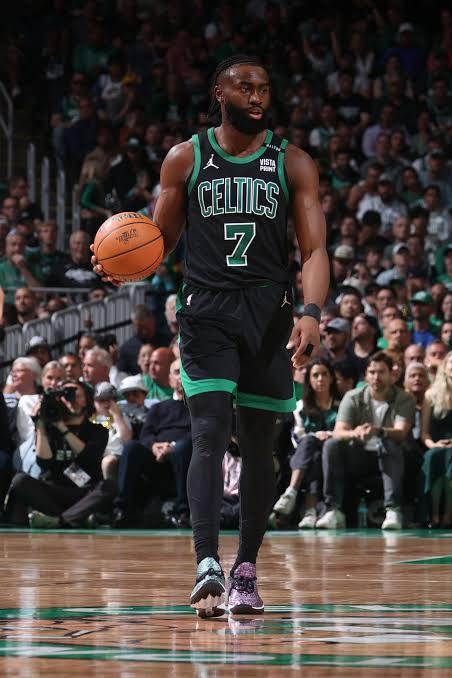 NBA player Jaylen Brown walking on the court holding a basketball, wearing a black Boston Celtics number 7 jersey, with fans seated in the background.