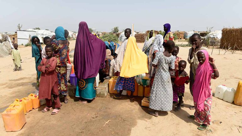 A group of children and women in a rural Northern Nigerian community queuing with yellow jerrycans to fetch water at a manual borehole.