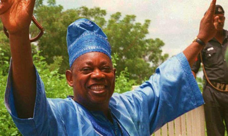 Close-up of a smiling Chief MKO Abiola raising both arms in celebration, wearing a blue traditional Yoruba agbada and cap, with greenery and a security official blurred in the background.