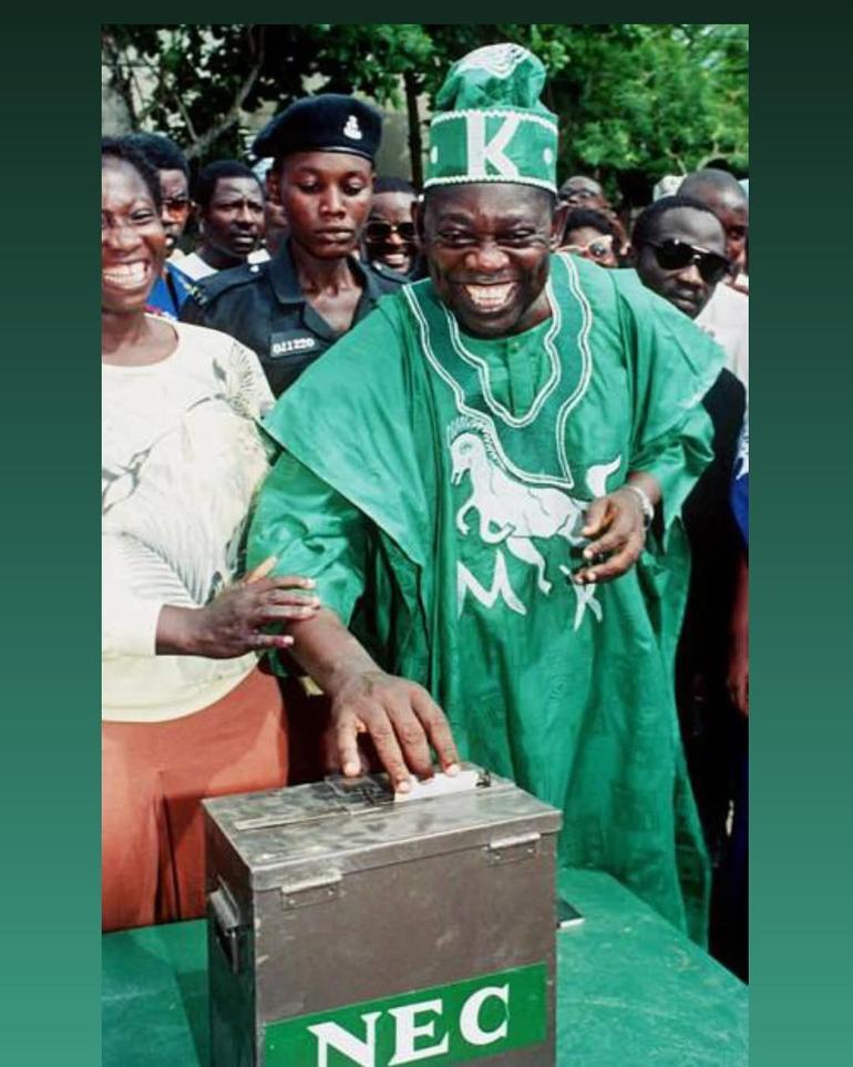 Chief MKO Abiola smiling while casting his vote in a metal NEC ballot box during the June 12, 1993 presidential election in Nigeria. He is wearing a green traditional Agbada and a matching cap with the letter 'K'.