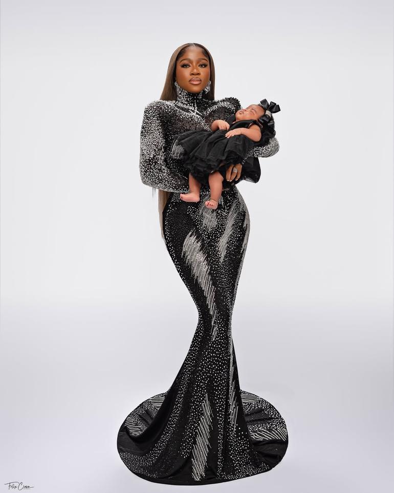 Full-length portrait of Veekee James in a sparkling black and silver mermaid dress holding her sleeping baby girl, Eliana Atere.