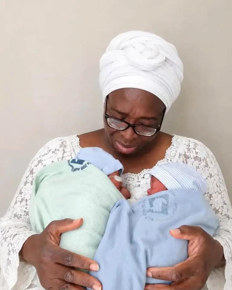 59-year-old Nigerian woman looks down emotionally while cradling her twin babies.