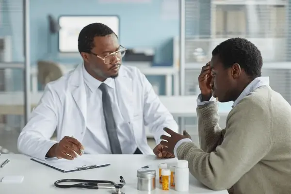 A middle-aged Black male doctor discussing health concerns with a young male patient in a bright clinic.