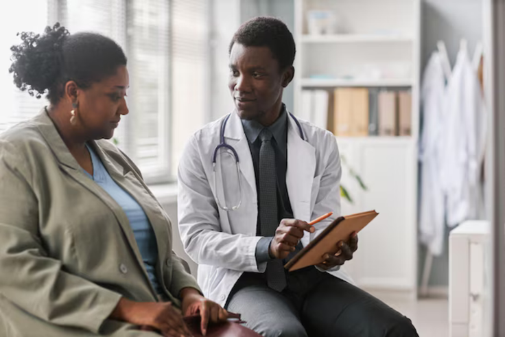 A male Nigerian doctor in a white coat showing medical data on a tablet to a female patient in a professional hospital setting.