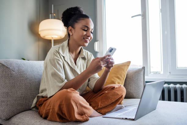 A smiling young woman sitting cross-legged on a couch in a bright living room, holding a smartphone and looking at the screen with a laptop open in front of her.