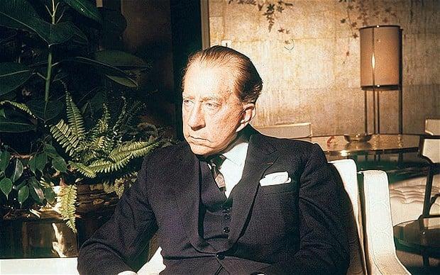 An older J. Paul Getty wearing a dark three-piece suit and pocket square while sitting in a modern room next to a leafy indoor plant.
