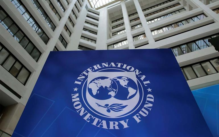 The International Monetary Fund (IMF) logo with its globe design on a large blue banner inside a building with a high atrium.