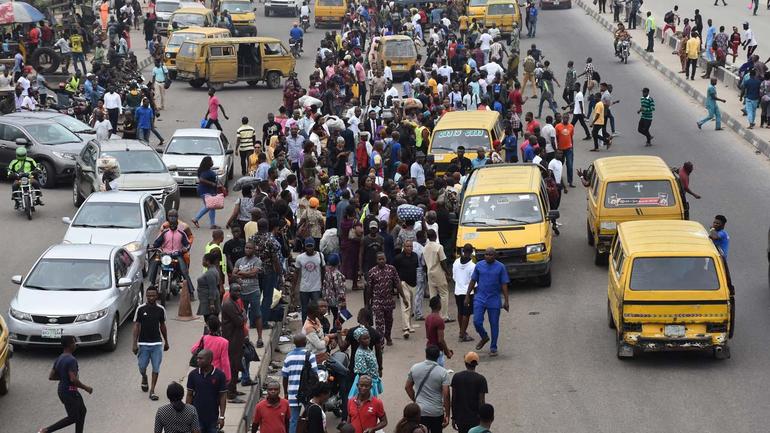 A busy Lagos street scene showing yellow commercial buses (danfos), private cars, and many pedestrians moving in traffic.