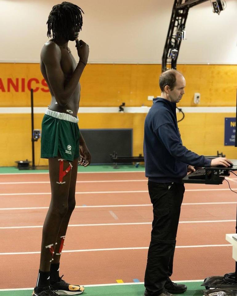 Side profile of 7'6" South Sudanese-Australian basketball recruit Jongkuch Mach standing next to a technician at a computer workstation during a biomechanics evaluation.