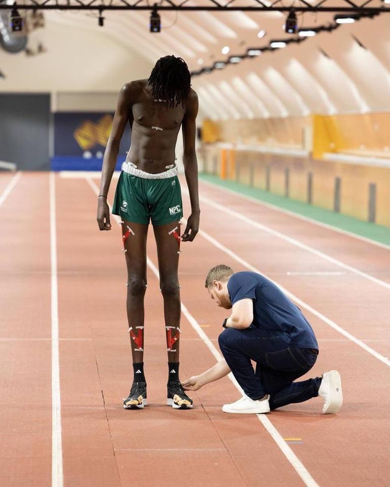 A sports scientist kneeling down on an indoor track to adjust motion-tracking sensors on the ankles of 7-foot-6 basketball player Jongkuch Mach.