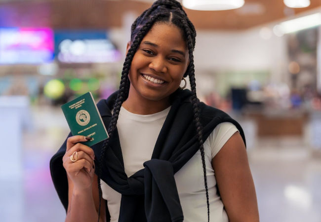 A smiling young woman holding up a green Nigerian passport inside a modern building.
