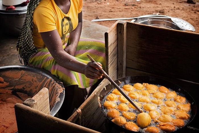 A woman sitting outdoors and frying golden-brown Akara balls in a large black pan filled with hot oil.