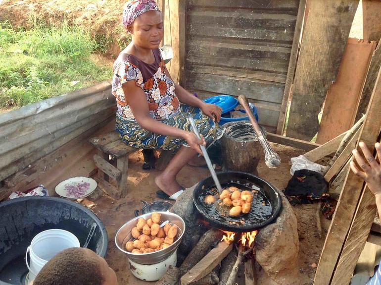 A woman cooking outdoors, stirring bean cakes over an open firewood flame with a bowl of finished Akara next to her.