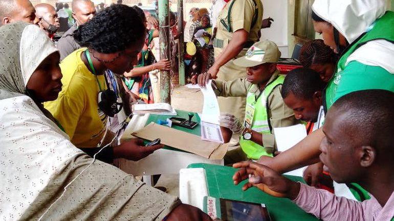 NYSC corps members in uniform and high-visibility vests working as INEC ad-hoc staff at a busy Nigerian polling unit during an election.